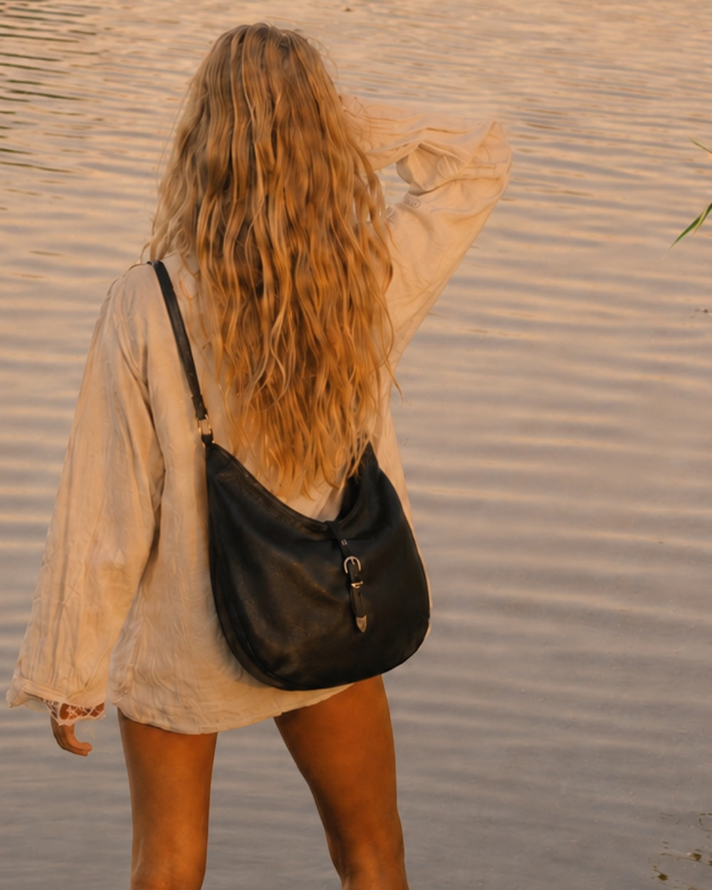 Urban Originals USA Lighthouse black pebbled vegan leather hobo bag with silver buckle worn by a woman at sunset